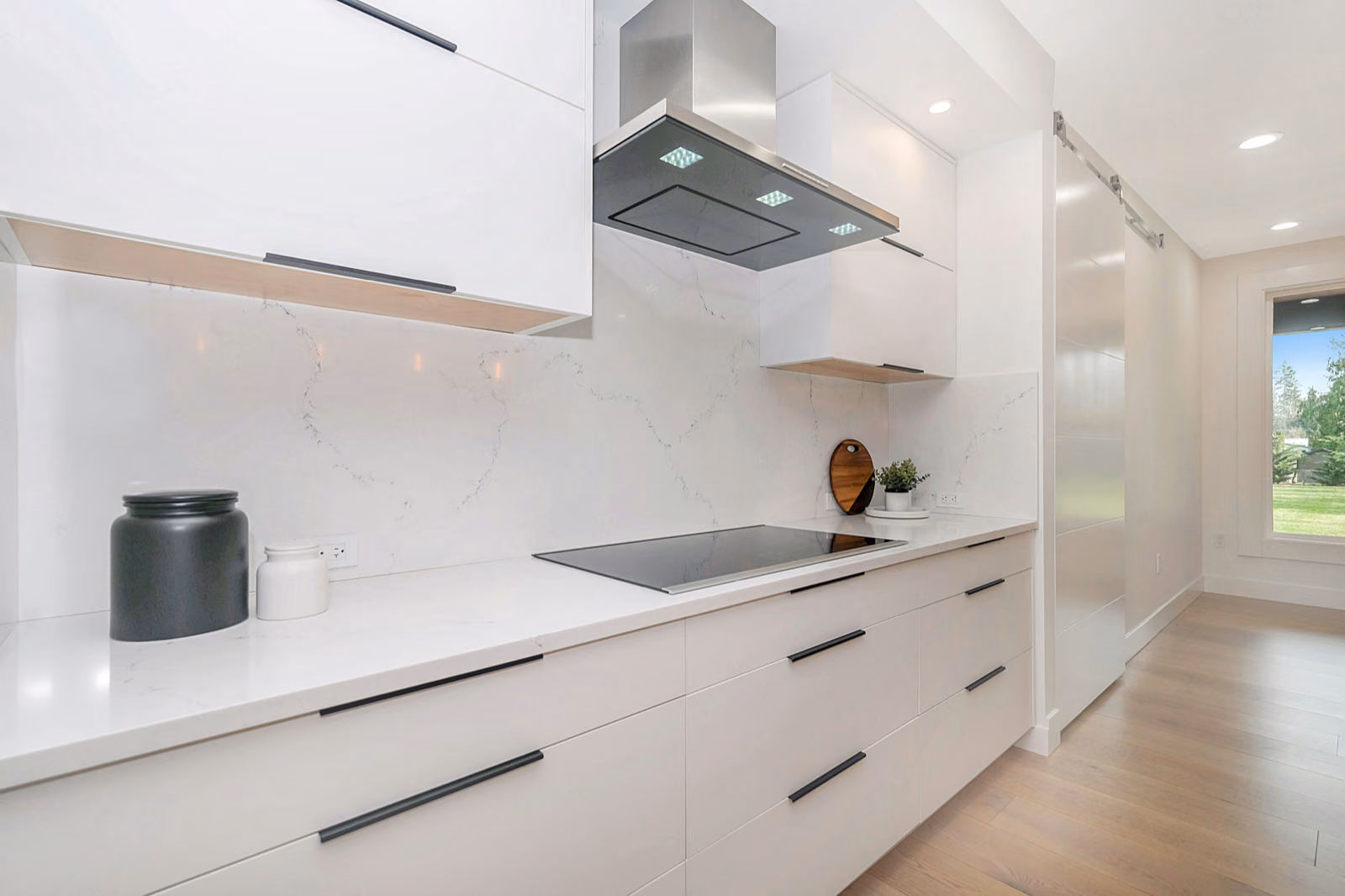 A bright, airy kitchen with Renotive high-end appliances, a large central island with a quartz countertop, and pendant lighting casting a warm glow. Wooden cabinets with a modern, minimalist design line the walls, complemented by a backsplash of natural stone tiles. The room is flooded with natural light from large windows overlooking a well-manicured backyard. Subtle pops of color, such as a vase of fresh flowers, add visual interest. The overall atmosphere is one of elegance, functionality, and a seamless blend of traditional and contemporary elements.